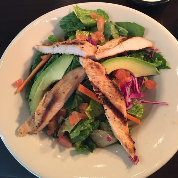 Grilled chicken salad with avocado, lettuce, tomatoes, and shredded cabbage in a white bowl.